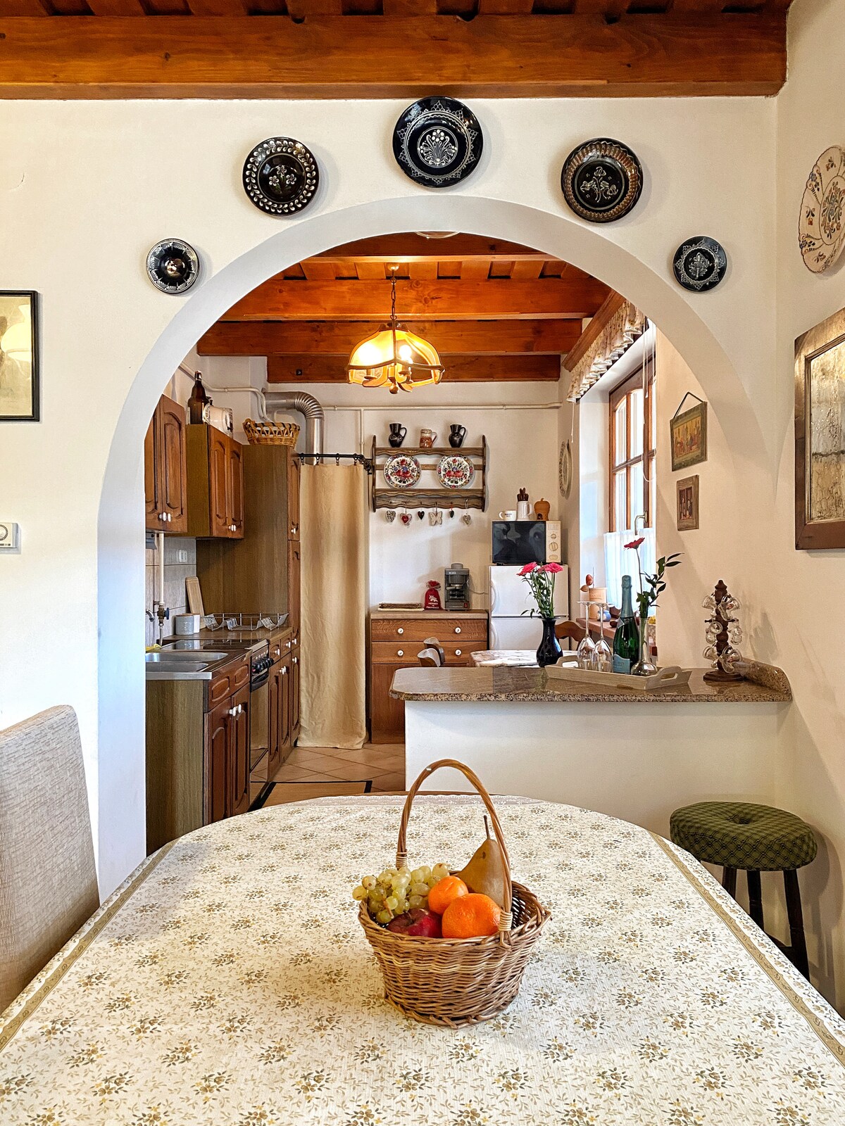 A cozy kitchen area features an archway leading to cooking spaces adorned with wooden cabinets and various decorative plates on the walls. A basket filled with fruits sits on a table covered with a patterned cloth, complemented by a rustic chandelier overhead.