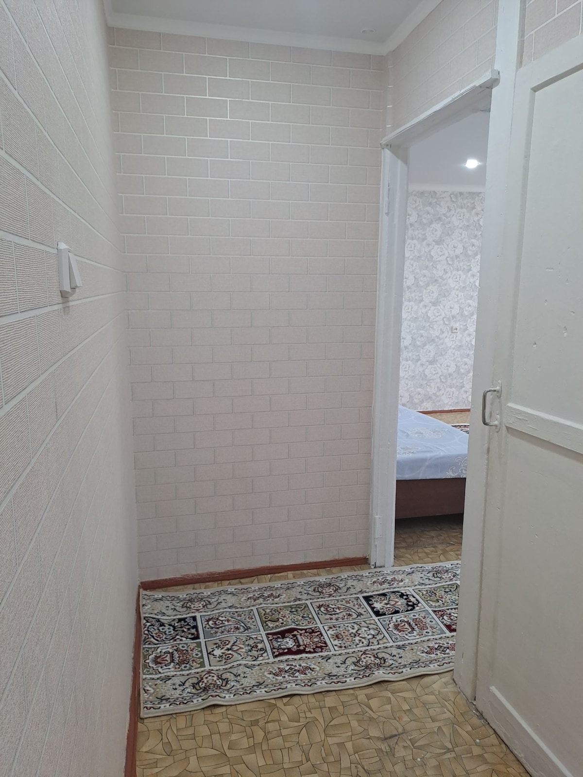 A narrow hallway displays light-colored, textured walls with a subtle pattern. A decorative area rug lies on the floor, adding warmth. A door is visible at the end of the hallway, leading to another room, while soft lighting illuminates the space.