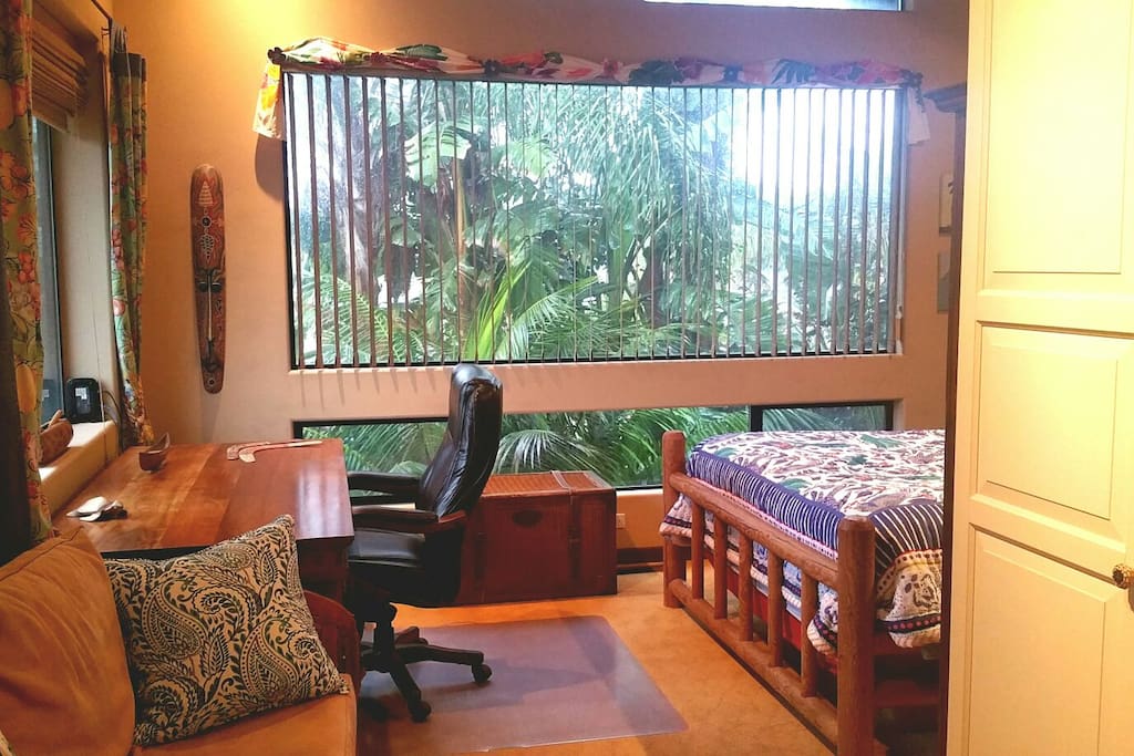 View from entry of generously-sized room. Large 4' x 10' picture window with blinds showing view of tropical palms and giant bird of paradise just outside. Tropical hardwood desk back left and sofa with wardrobe/TV in foreground.