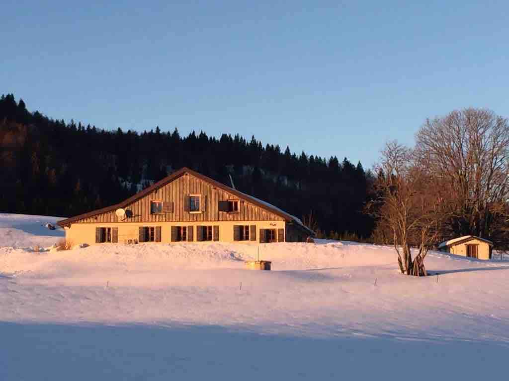 A rustic farmhouse is set against a snowy landscape just before sunset. The structure features a sloped roof, multiple large windows, and is surrounded by open fields. A small outbuilding is visible to the right, framed by bare trees in a serene, winter setting.