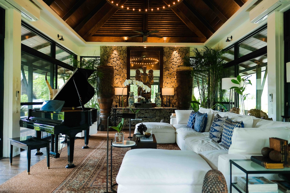 A spacious living area features a grand piano positioned next to large glass windows, allowing natural light to fill the room. Comfortable white sofas are adorned with decorative pillows. A central stone wall and several plants enhance the tranquil atmosphere of the space.