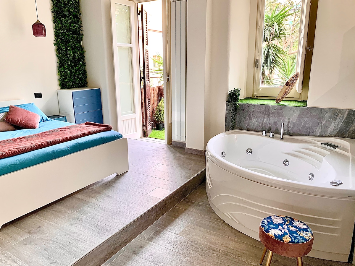 An airy open space features a white, spacious bed with blue and red accents. Adjacent to it, a round hot tub invites relaxation, with greenery visible through the open doors. Natural light floods the room, illuminating the warm wood flooring.