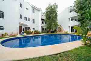 A serene outdoor area features a large, oval-shaped pool surrounded by lush greenery. The edges are lined with smooth stone, providing a space for relaxation. Nearby buildings, designed with white facades and decorative windows, stand in a peaceful arrangement, contributing to a tranquil atmosphere.