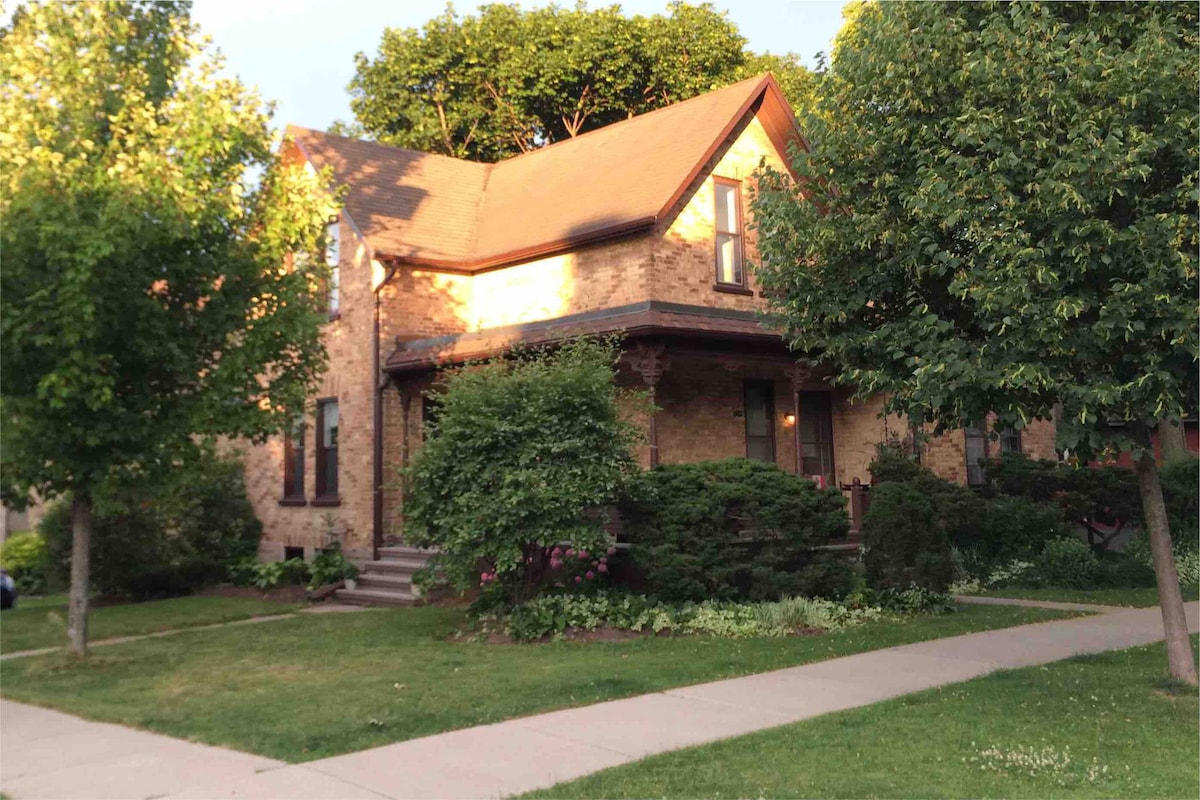 A charming brick building with a sloped roof is surrounded by greenery and well-maintained landscaping. The entrance is flanked by shrubs and small trees, creating a welcoming outdoor space. A paved sidewalk leads to the front door, enhancing accessibility to the home.