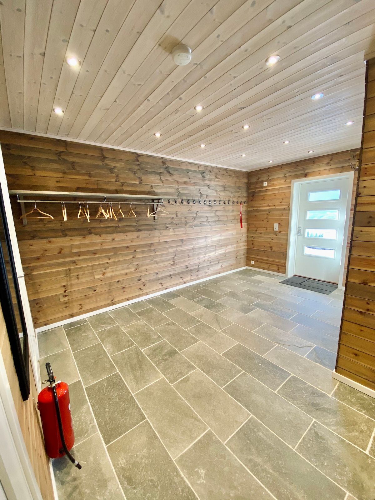 An entryway features wooden paneling and modern lighting fixtures on the ceiling. A stone floor provides a durable surface, while a coat rack and pegs line one wall for storage. The glass door allows natural light to enter, creating an open atmosphere.