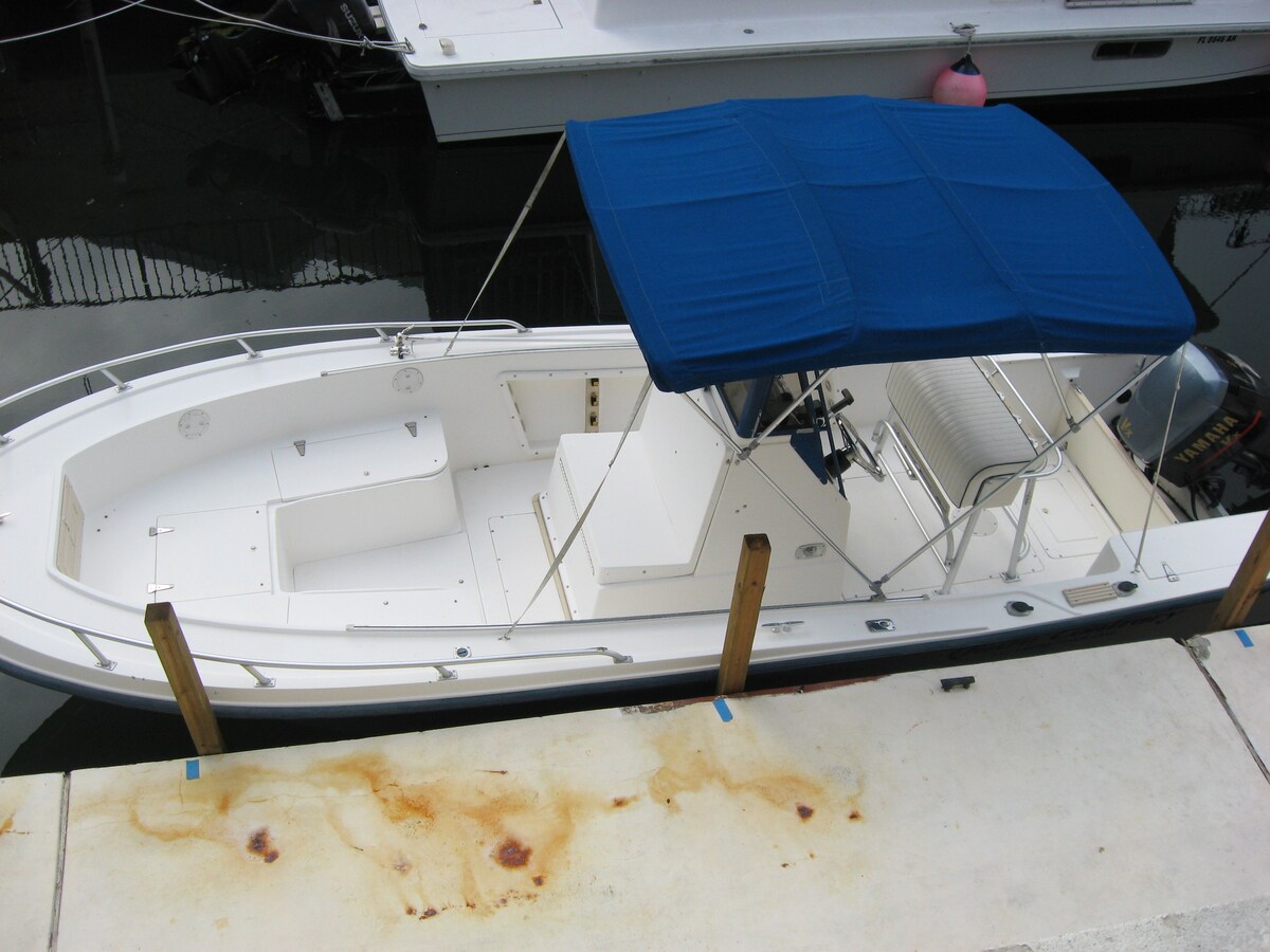 A center console boat is docked securely at a marina, featuring a blue canopy providing shade. The boat's spacious interior includes seating and ample deck space. The surrounding area displays a calm waterway, with several other boats visible in the background.