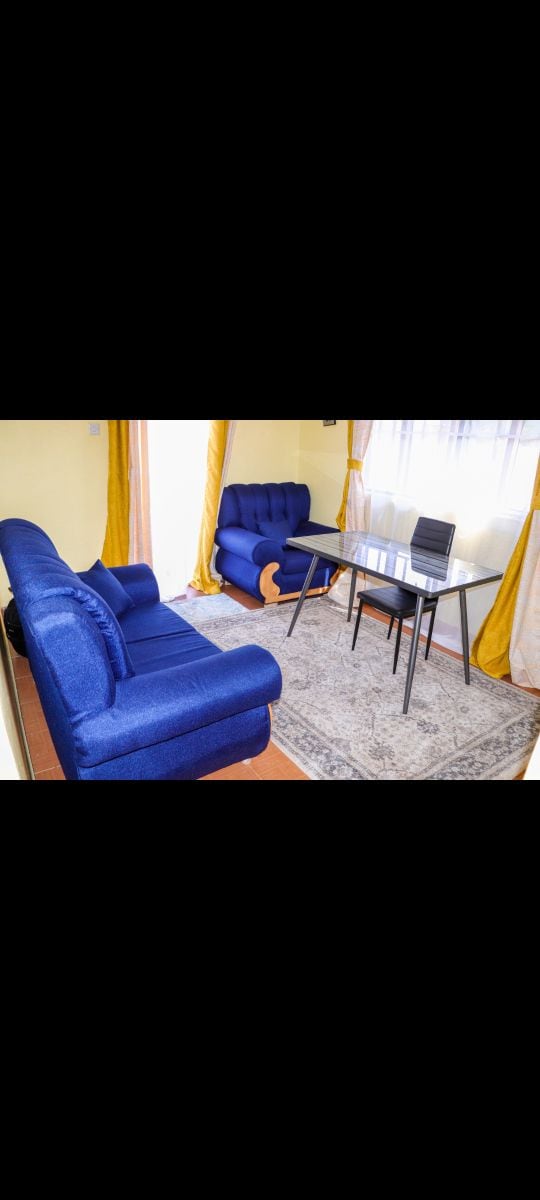 A living area features a bright blue sofa and armchair, arranged around a glass table with a black chair. Natural light enters through sheer curtains, enhancing the warm yellow walls and soft area rug underfoot.