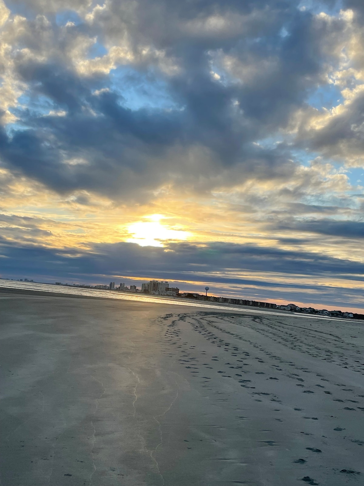 The image features a beach scene at sunset, with footprints etched in the wet sand leading towards the horizon. Dramatic clouds cast varying shades across the sky, mixing golden sunlight with soft blues and grays. A distant skyline is visible along the shore.