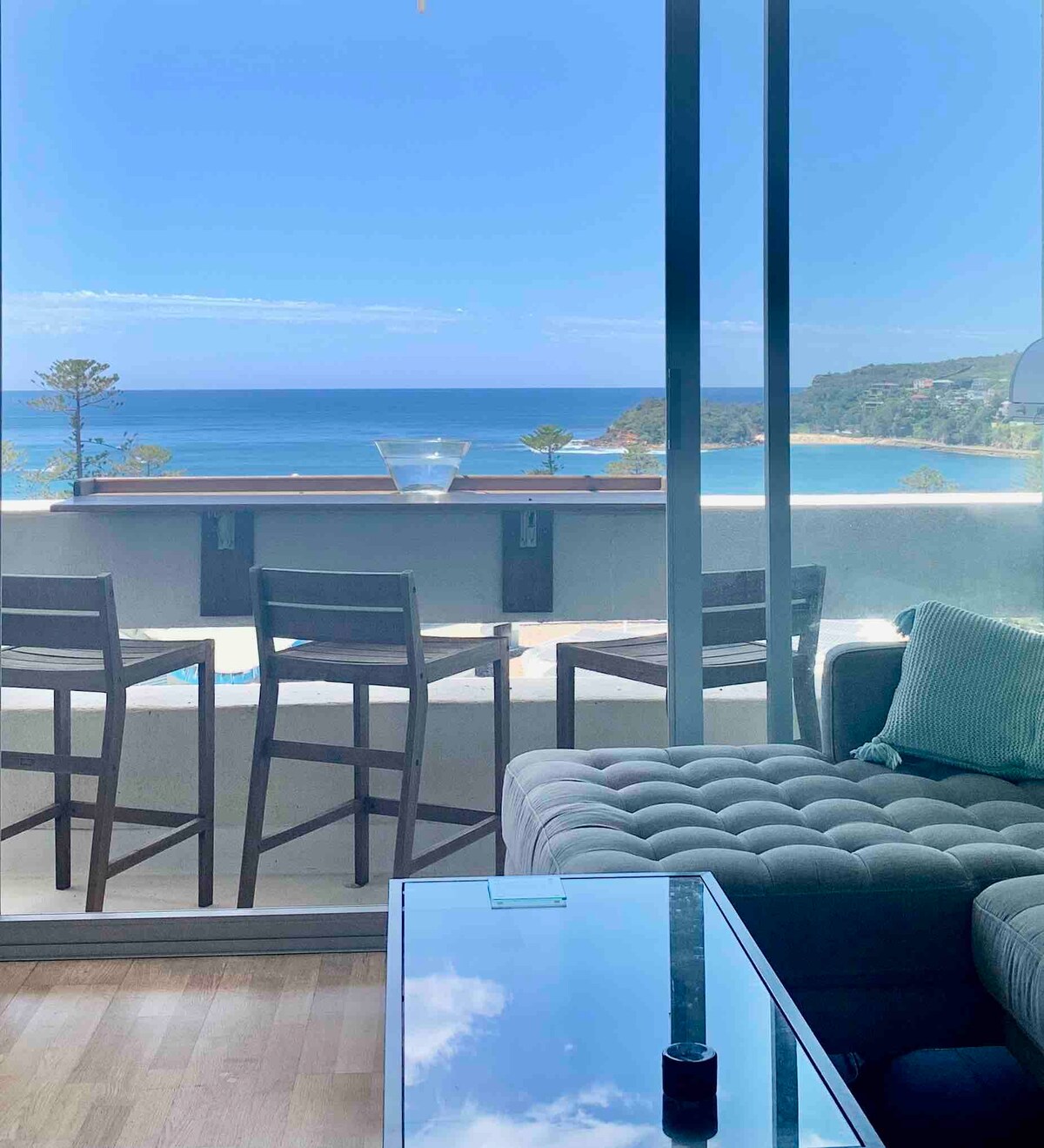 The living room features a modern lounging area with a comfortable sofa and a glass coffee table. Large windows provide expansive ocean views, while a private balcony with four high-backed bar chairs overlooks the water, creating a seamless connection to the stunning coastal scenery.