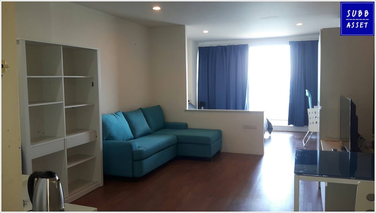 A spacious living area features a turquoise sectional sofa and a modern desk positioned near a large window with navy blue curtains. A bookshelf and a kettle are also visible, creating a functional and inviting space.