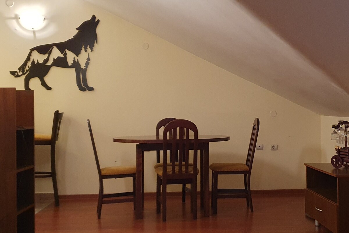 A dining area is presented with a wooden table surrounded by four matching chairs. A black wall decoration of a wolf silhouette is displayed prominently, while soft lighting enhances the cozy feel of the space.