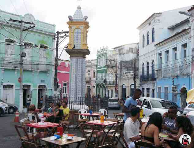 A lively street scene is depicted, featuring colorful buildings in shades of pink, green, and blue. Small tables with chairs are arranged outside, where individuals can be seen dining. A prominent tower, adorned with decorative elements, stands in the center of the square, framed by a wrought iron fence.