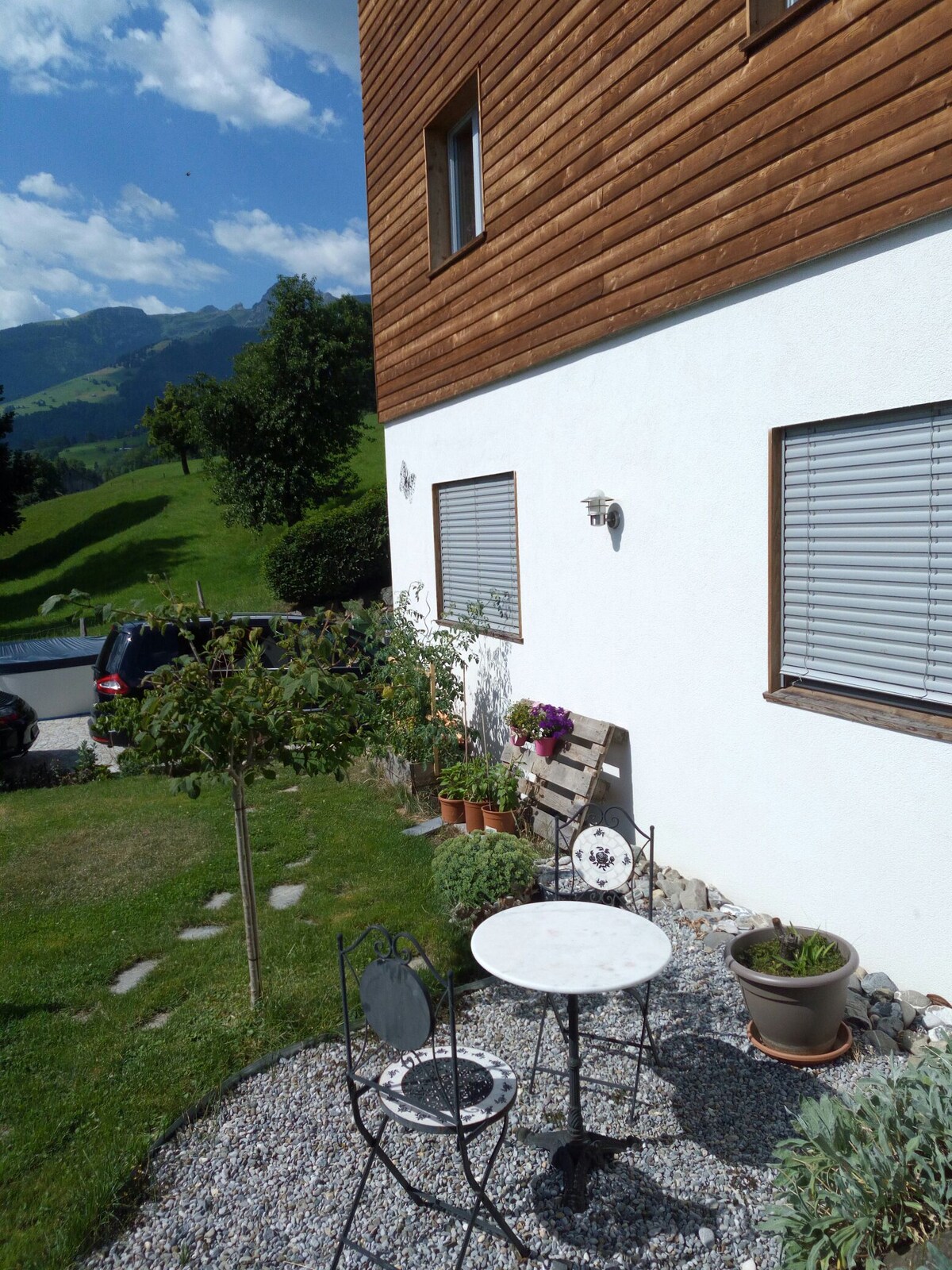 A charming outdoor seating area is depicted, featuring a small round table with two chairs, surrounded by greenery and flower pots. The building's wooden siding complements the peaceful landscape, with mountains visible in the distance under a blue sky.