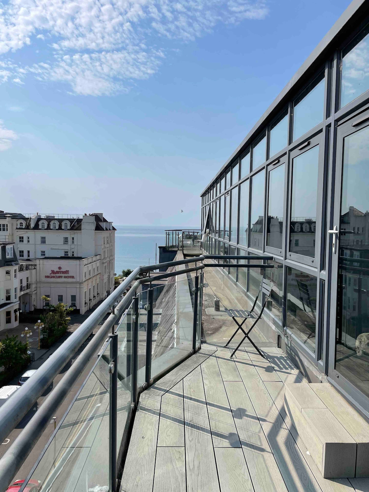 A spacious balcony extends from the penthouse, featuring glass panels that provide unobstructed views of the sea. A single chair is positioned near the edge, offering a serene spot to enjoy ocean breezes and sunlight. The deck is finished with wooden planks, enhancing the outdoor experience.