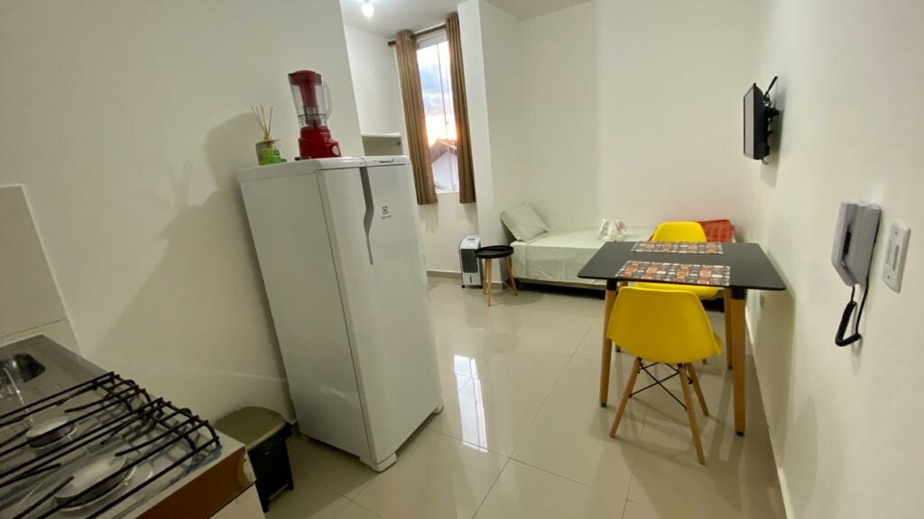 A bright kitchen area features a compact refrigerator and a cooking range alongside a small dining table with yellow chairs. A light green seating option is positioned near a window, while a mounted television is installed on the wall. Natural light fills the space.