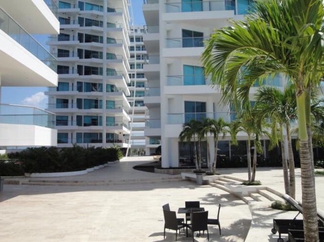 A modern architectural complex is surrounded by palm trees and landscaped areas. The image highlights the use of glass and concrete in the buildings, with outdoor seating arrangements visible on a spacious terrace. Sunlight enhances the clean lines and open space.