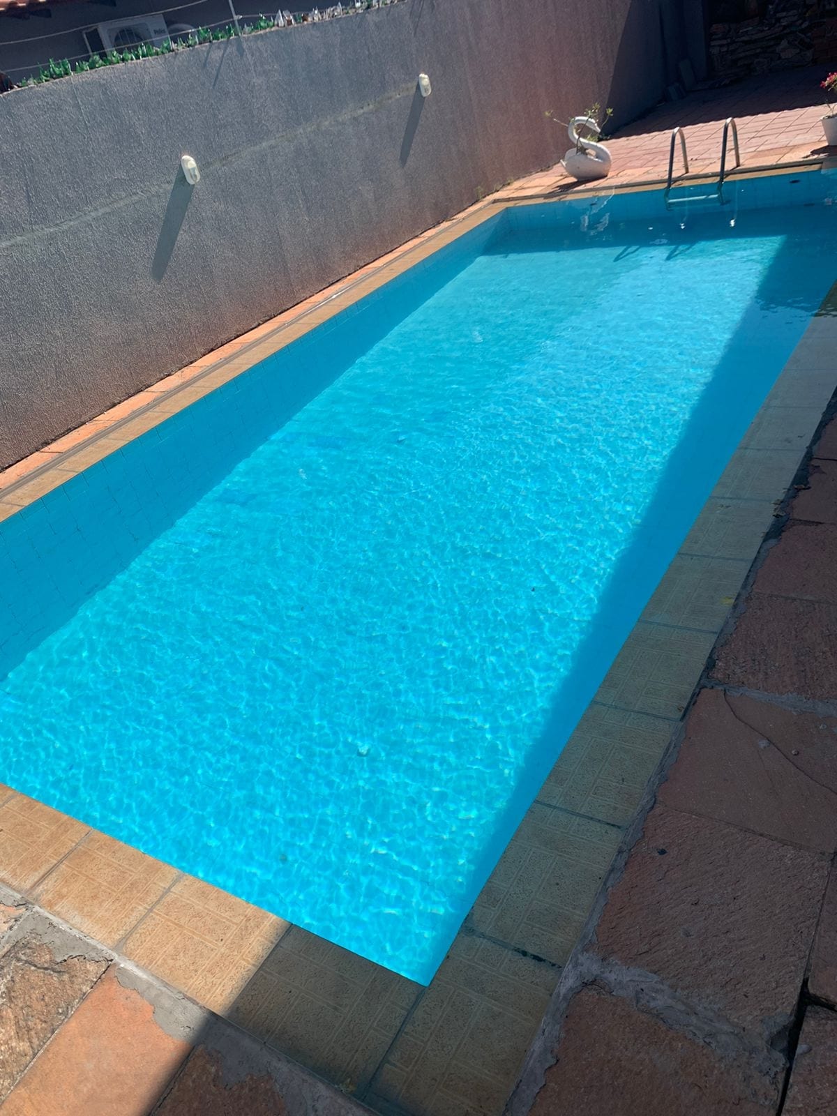 A private pool is presented, featuring clear blue water that reflects sunlight. The surrounding area is bordered by a stone deck, providing a spacious feel. A small potted plant adds a touch of greenery beside the pool's edge, enhancing the tranquil environment.