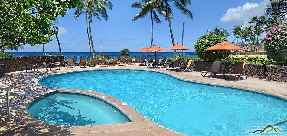 A heated oceanfront pool is surrounded by lounge chairs, with orange umbrellas providing shade. Lush greenery and palm trees are visible, framing the serene view of the ocean and sky. The pool area offers a welcoming space for relaxation and enjoyment.