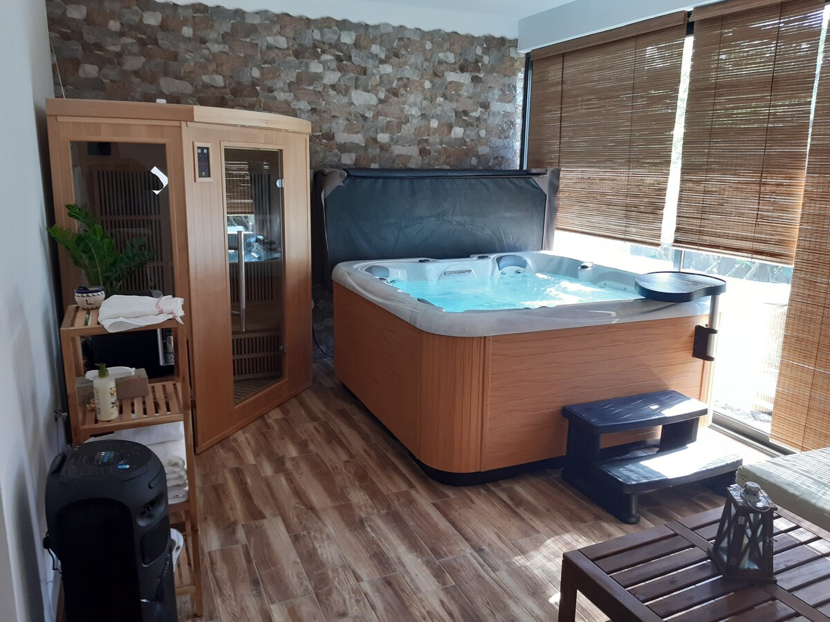 A wellness space features a hot tub alongside an infrared sauna, both framed by a natural stone wall. Bamboo blinds filter natural light, while a small wooden shelf holds towels and additional amenities. A cozy seating area completes this inviting relaxation space.