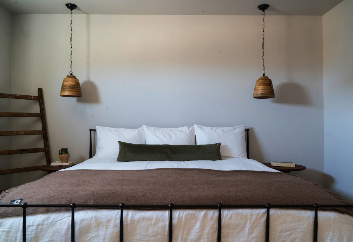 A spacious bed is center stage in the image, dressed in neutral linens and accented by a brown throw blanket. Two pendant lights hang above the bed, and a simple wooden ladder is positioned to the left, adding a touch of rustic charm.