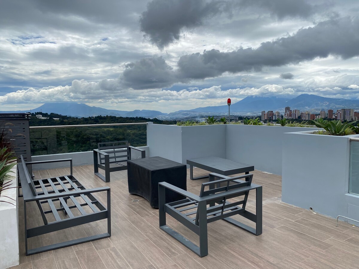 A rooftop terrace is furnished with several modern seating arrangements, including four lounge chairs and two side tables. The area offers sweeping views of the surrounding mountains and city skyline, complemented by a railing for safety and greenery along the edges.