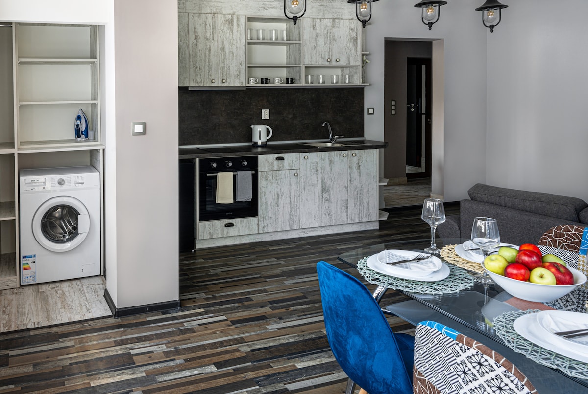 A modern kitchen and dining area are visible, featuring light wood cabinetry and a countertop with a stove. A washer is positioned nearby. A glass dining table set for four sits in the foreground, complemented by colorful fruit in a bowl. Soft lighting is provided by fixtures above.