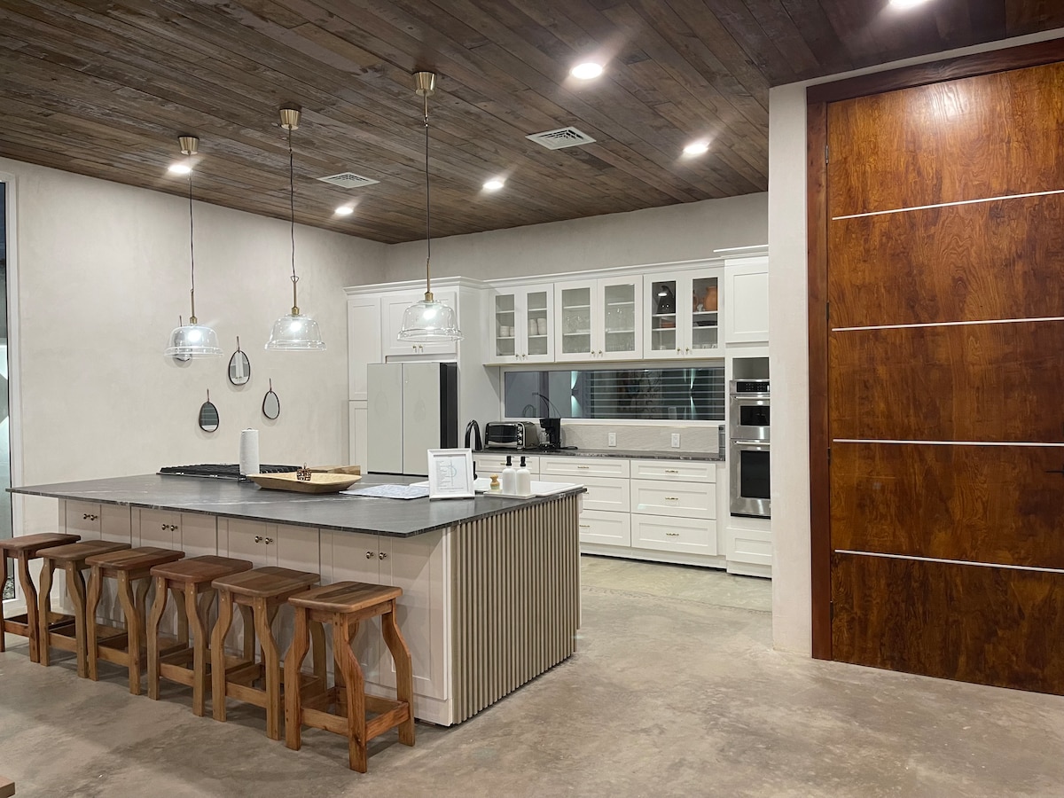 A modern kitchen features a large island with seating for six, made of natural materials and complemented by wooden bar stools. White cabinetry and appliances enhance the bright, open space, while the ceiling showcases rustic wooden beams, contributing to a warm yet contemporary ambiance.