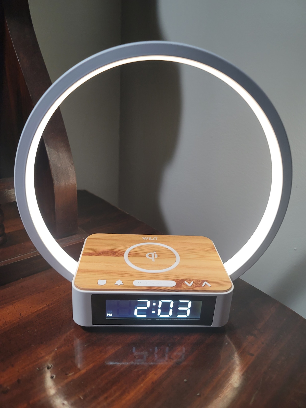 A sleek alarm clock with a circular lamp design is displayed on a polished wooden surface. The clock features a digital display showing the time, with a soft glow emanating from the light ring, creating a contemporary appearance.