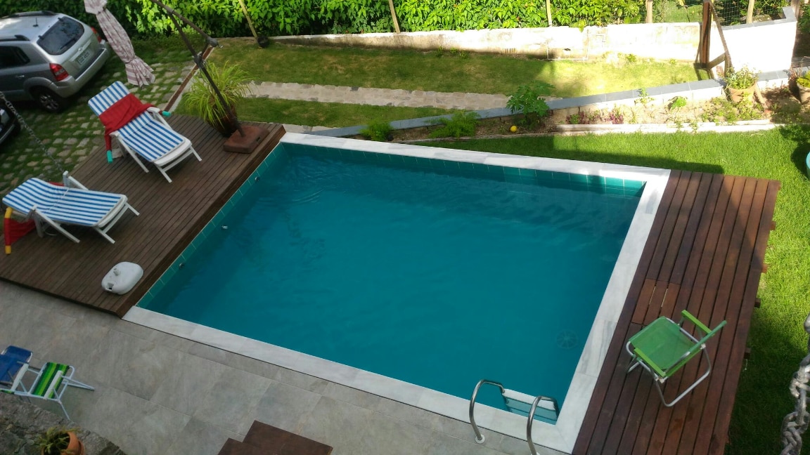 A swimming pool is situated in a landscaped outdoor area, surrounded by sun loungers and a grassy expanse. The pool is framed by wooden decking and features a ladder for entry. Lush greenery and garden elements are visible in the background.