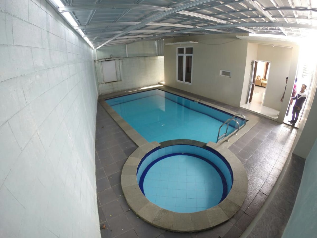 An indoor pool area features a large swimming pool alongside a circular hot tub. The walls are finished in light colors, and natural light filters in from a window, creating a bright and inviting environment. Steps lead into the pool, enhancing accessibility.