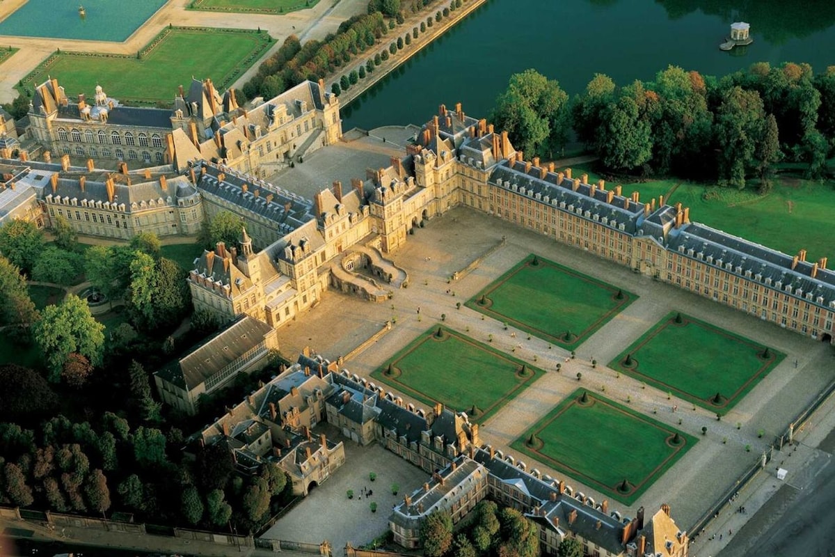 An aerial view captures a grand historical building surrounded by manicured lawns and trees. The symmetrical layout of the structure and nearby gardens reflects classical architecture. A shimmering body of water is seen in the background, enhancing the scenic beauty of the estate.