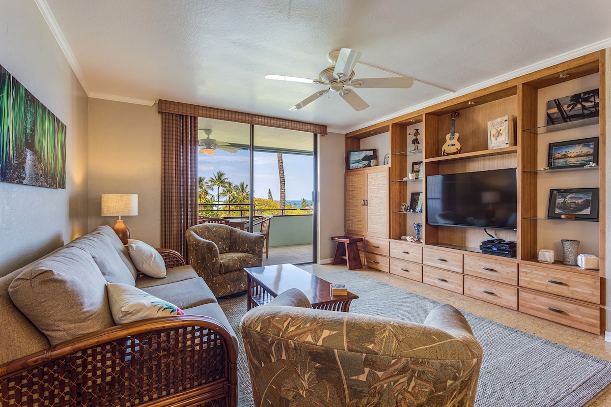 A bright living area is furnished with a comfortable sofa and two patterned chairs. A wooden coffee table sits in the center, and a TV is mounted on a wooden media unit. Large windows allow for natural light and views of palm trees outside.