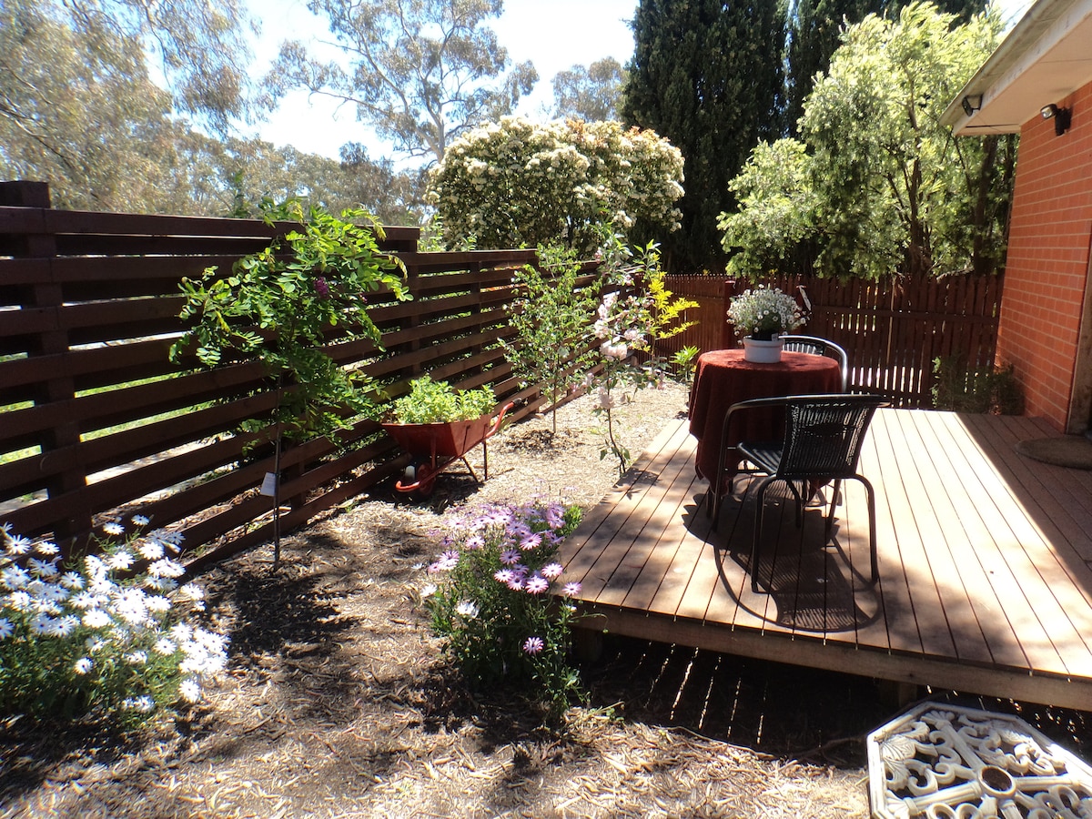 A private deck area is showcased with a round table and chairs, surrounded by lush flowers and greenery. A wheelbarrow and various plants add to the natural ambiance. The space is sunlit, providing a relaxing outdoor retreat.