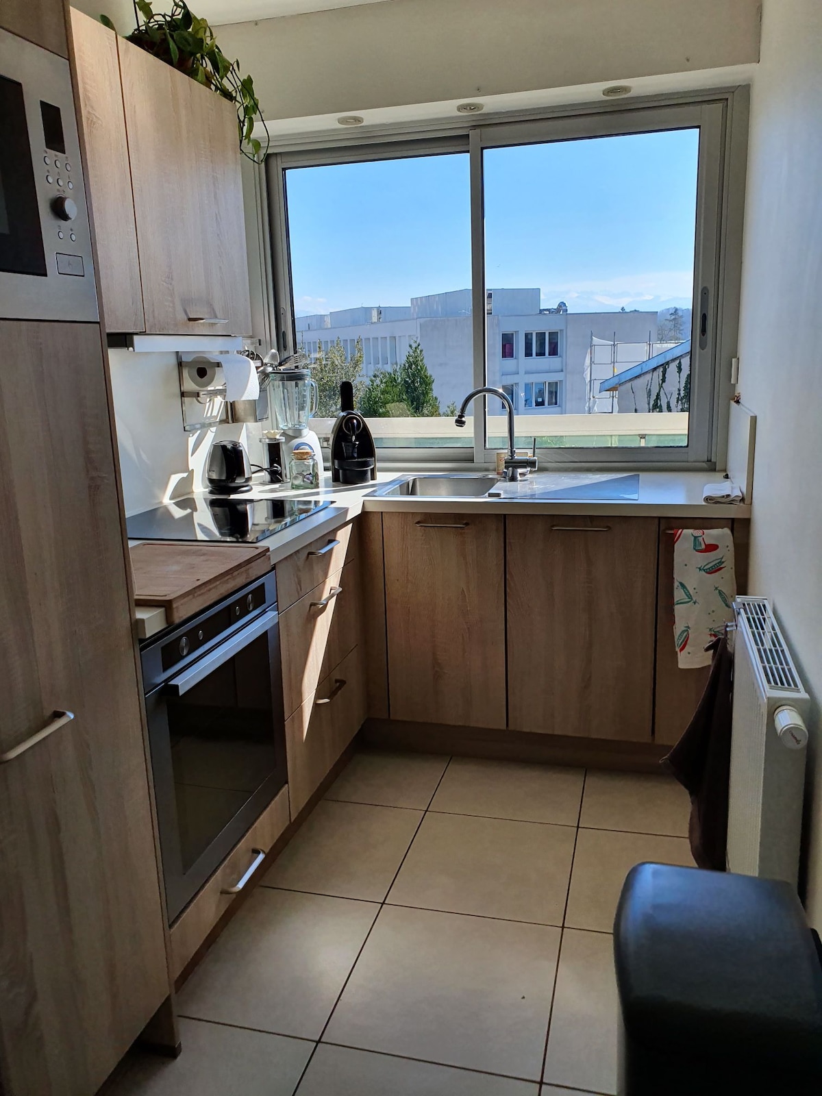 A compact kitchen features light wood cabinetry and modern stainless steel appliances. A window allows natural light to fill the space, offering a view of the outside. Counter space is utilized for a coffee maker and kitchen utensils, with tile flooring adding a clean finish.