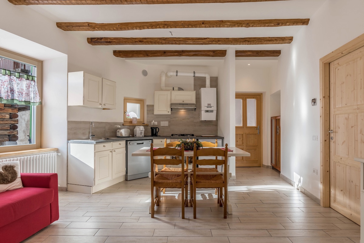 A well-lit kitchen is showcased with natural wooden beams overhead. Cream-colored cabinetry, a stove, and a refrigerator are included, along with a dining table surrounded by wooden chairs. A red sofa is visible in the corner, enhancing the cozy ambiance of the space.