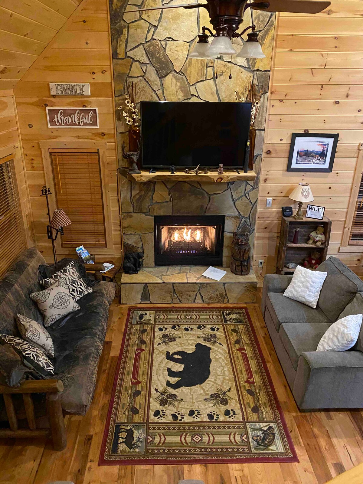 An open living space features a stone fireplace with a flickering flame, flanked by a comfortable gray couch and a rustic sofa. A patterned area rug with a bear motif rests beneath a coffee table, while a flat-screen TV is mounted above the fireplace.