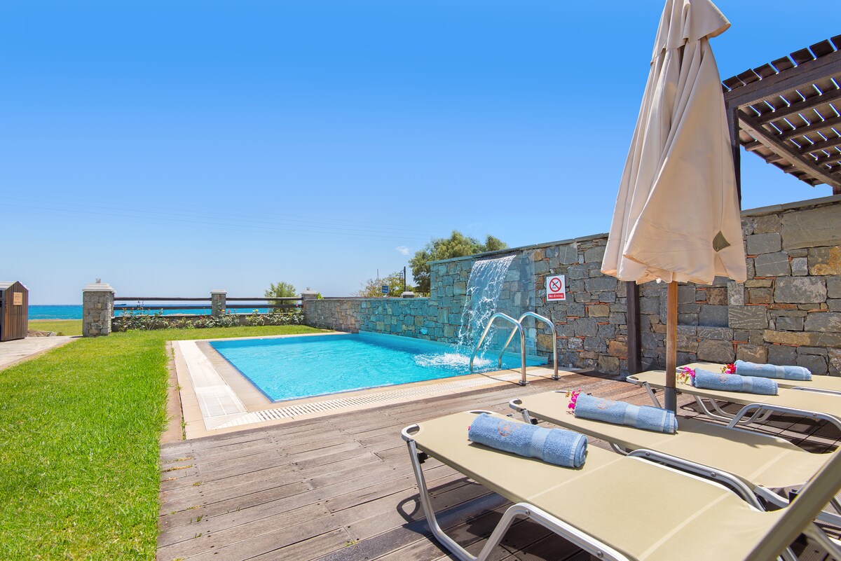A serene outdoor area is highlighted by a private pool featuring a gentle waterfall. Two sun loungers are positioned beside the pool, each neatly arranged with soft towels. Lush green grass surrounds the space, enhancing the overall relaxation setting.