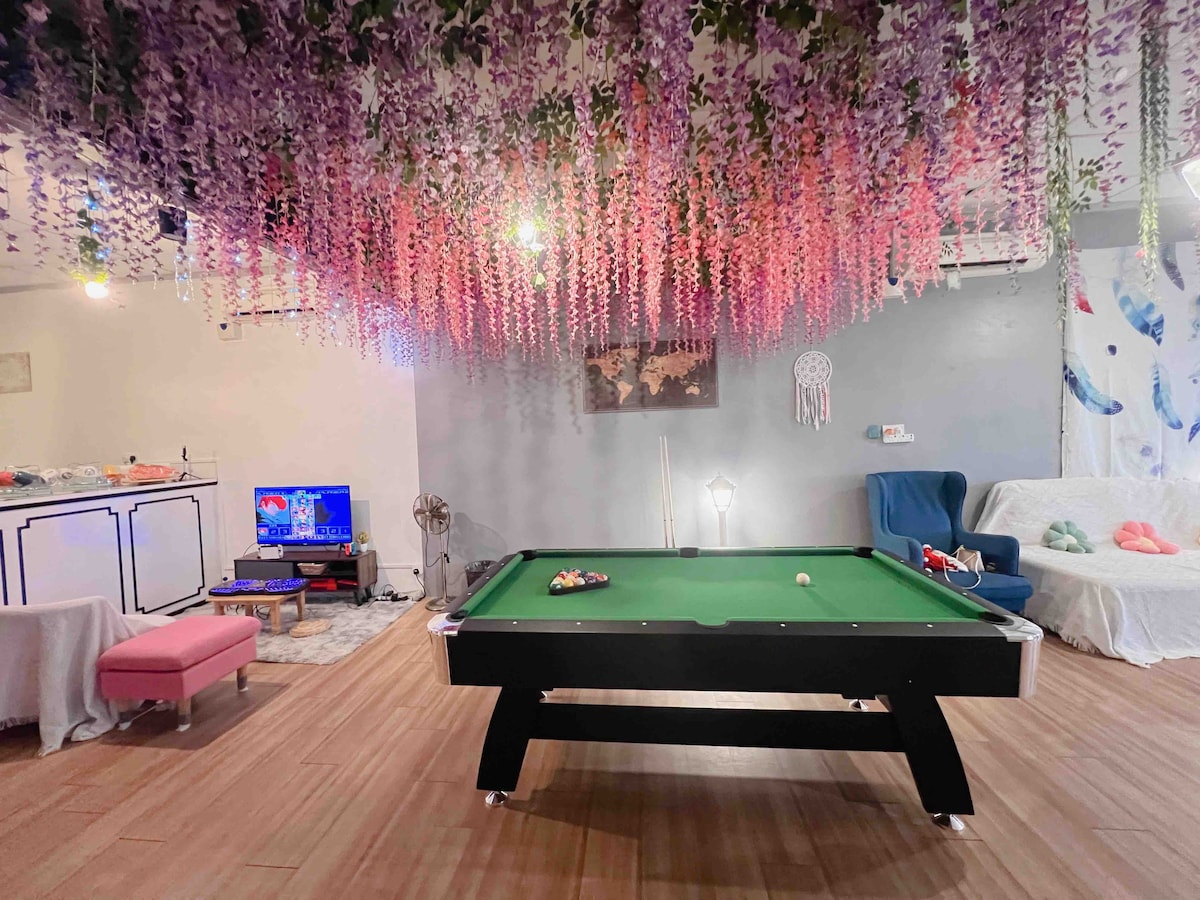 A game room is shown, featuring a sleek pool table at the center, surrounded by casual seating. Decorative floral accents hang from the ceiling, creating a vibrant atmosphere. A television is positioned nearby, enhancing the entertainment options available in the space.