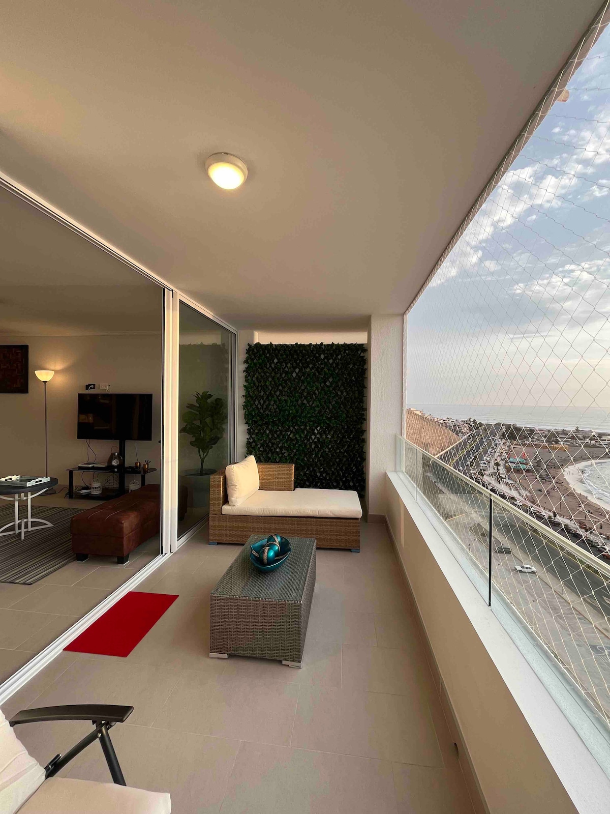 A sunlit balcony features a comfortable seating area with a lounge chair and a small coffee table. Greenery adorns one wall, adding a touch of nature. The scenic views of the coastline are framed by large glass sliding doors that enhance natural light.