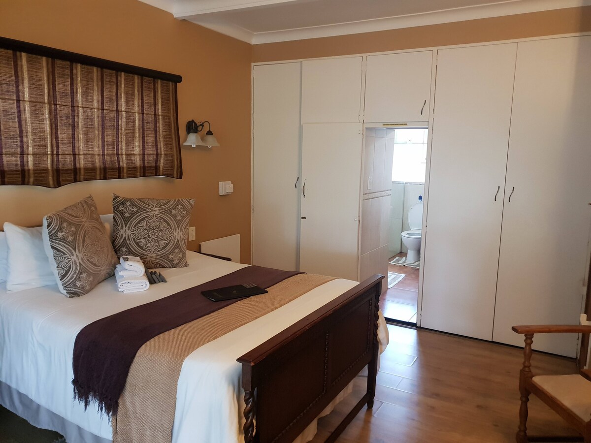 A well-appointed guest room features a spacious bed with a dark throw blanket and decorative cushions. Light floods in through a window dressed with textured curtains. Built-in white wardrobes are visible, and a private bathroom can be seen through an adjoining door.