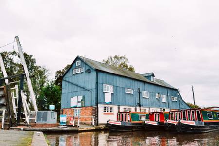 A blue-clad building with multiple windows stands adjacent to a calm waterway. Colorful boats are moored along the bank, reflecting the structure. The area is framed by trees and an adjacent loading crane, contributing to a serene waterside environment.