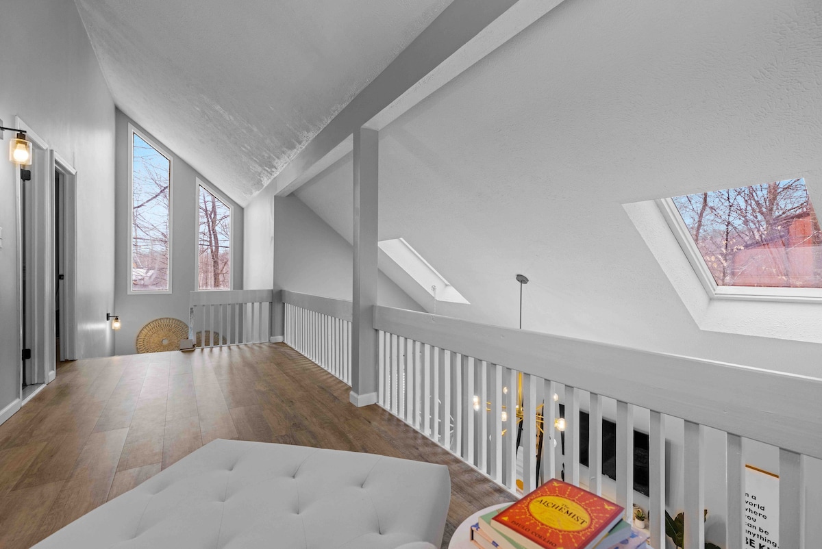 An airy loft space features sloped ceilings and large skylights, allowing natural light to fill the area. The wooden flooring complements a white railing, with a seating area and a stack of colorful books situated on a table. Calm and inviting vibes are present.
