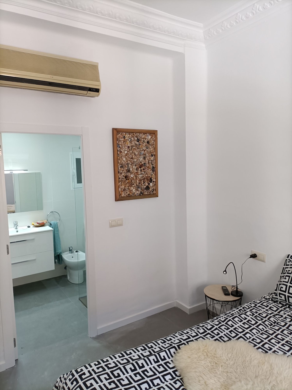 A bright bedroom corner showcases a stylish patterned bedspread on a spacious bed. A small bedside table holds a reading lamp, and a decorative wall art piece adds character to the space. An open door reveals a clean and modern bathroom area.