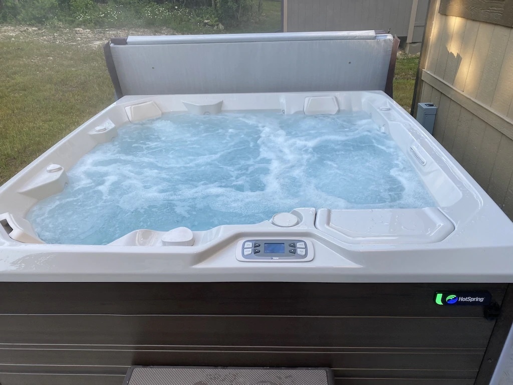 A spacious outdoor hot tub is filled with bubbling water, inviting relaxation. The control panel is visible, positioned on the side for easy access. Surrounding greenery and a glimpse of a nearby building add to the tranquil environment.