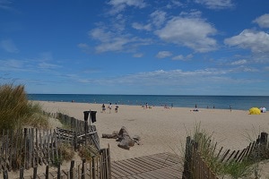A sandy beach is viewed from a wooden walkway. Waves lap gently at the shore, and a clear blue sky is dotted with fluffy white clouds. Beachgoers can be seen in the distance, enjoying the sun, while a yellow beach umbrella is visible on the sand.