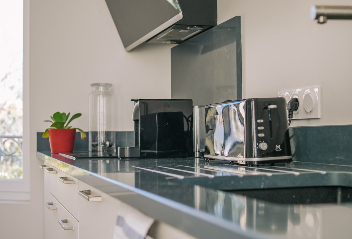 A modern kitchen countertop is presented, featuring sleek appliances including a coffee maker and a toaster. A small potted plant adds a touch of greenery, while ample natural light illuminates the space. The overall design emphasizes simplicity and functionality.
