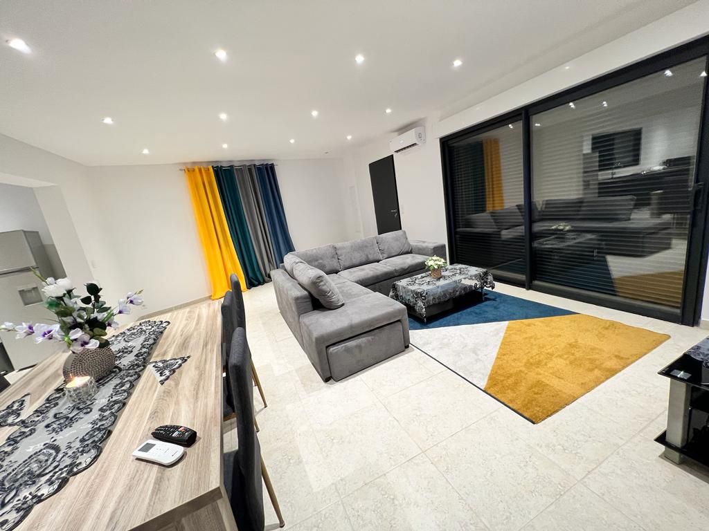 The living area features a spacious gray sectional sofa positioned near a patterned area rug. A wooden dining table is visible, adorned with a vase of flowers. Large glass doors allow natural light to enter and provide views of the outdoor space.