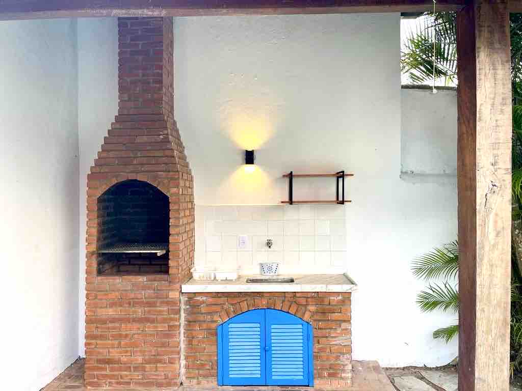 An outdoor grilling area features a brick barbecue beside a counter with a sink. The space is framed by wooden posts, and a bright blue cabinet adds a pop of color. A wall-mounted light fixture provides illumination, enhancing the functional outdoor space.