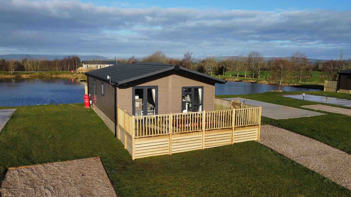 A luxury lodge is shown with a wooden exterior, surrounded by grassy areas and positioned near a tranquil lake. A deck with railings wraps around the lodge, providing outdoor space. Large windows offer views of the landscape, allowing natural light into the interior.