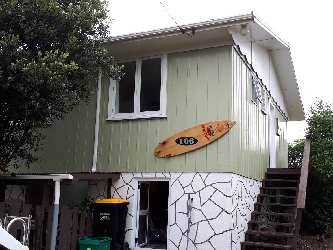 A beach house exterior is shown, featuring a light green upper level with large windows. A surfboard is mounted on the wall, displaying the number '106.' The ground level has a textured stone finish, and a staircase leads to the entrance.